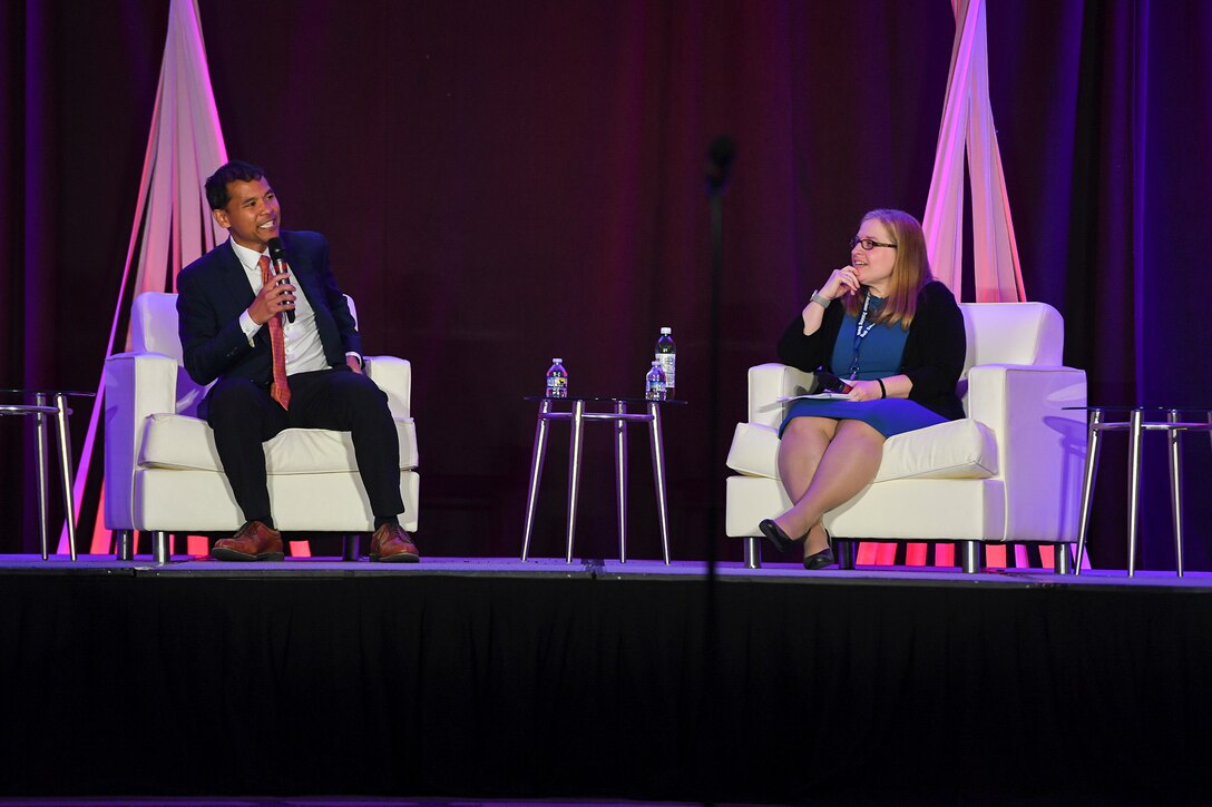 A man and woman wearing business attire sit across from one another on a stage.