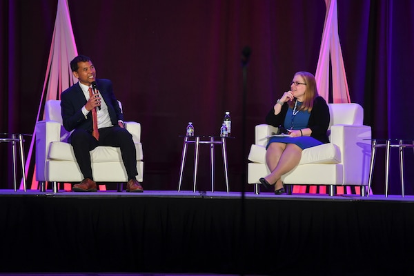 A man and woman wearing business attire sit across from one another on a stage.