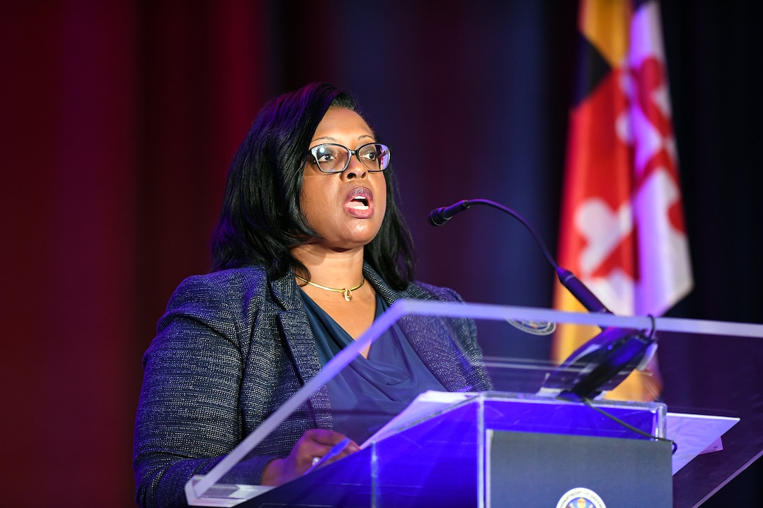 A woman wearing business attire speaks into a microphone while standing behind a lectern.