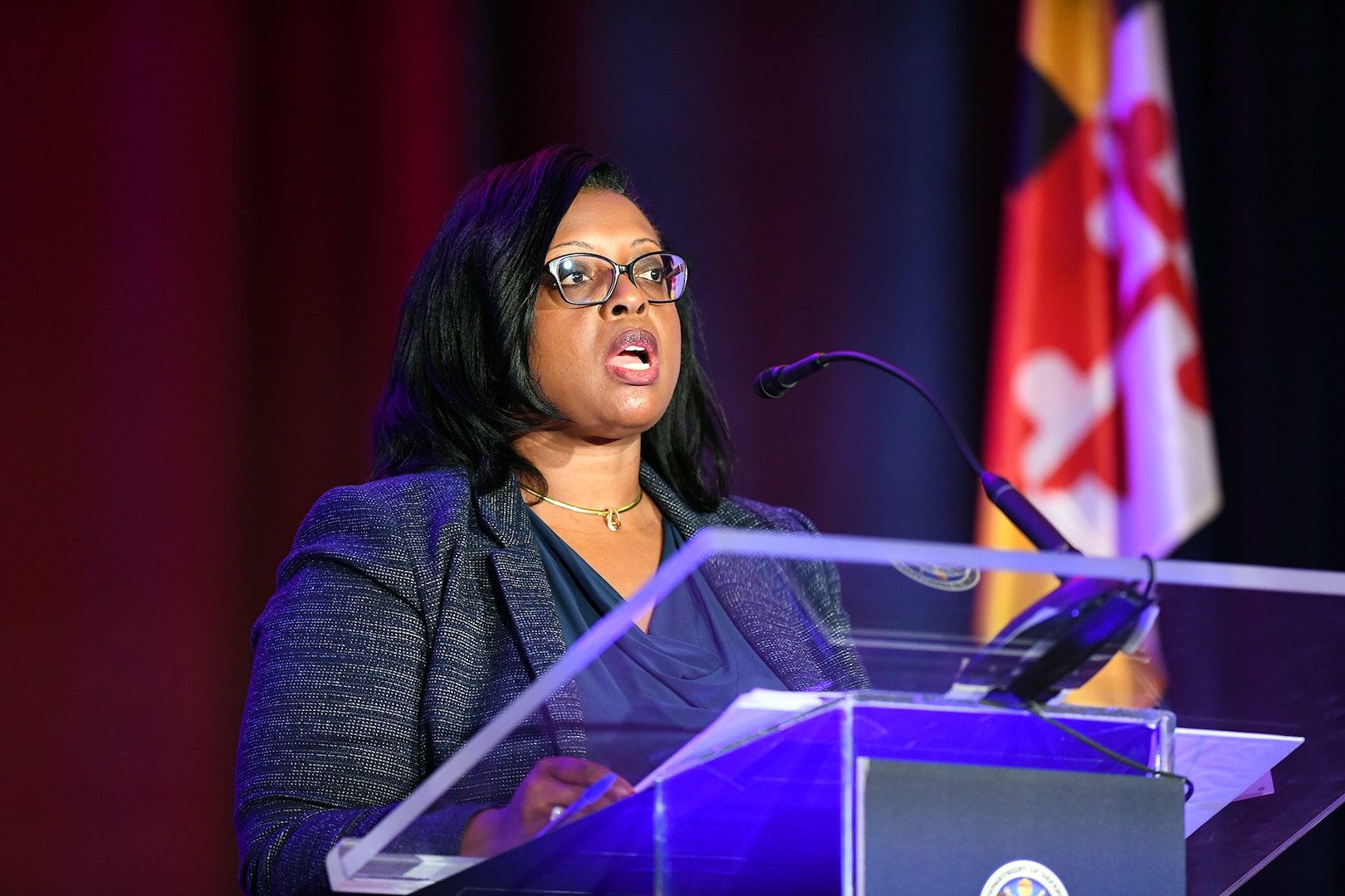 A woman wearing business attire speaks into a microphone while standing behind a lectern.