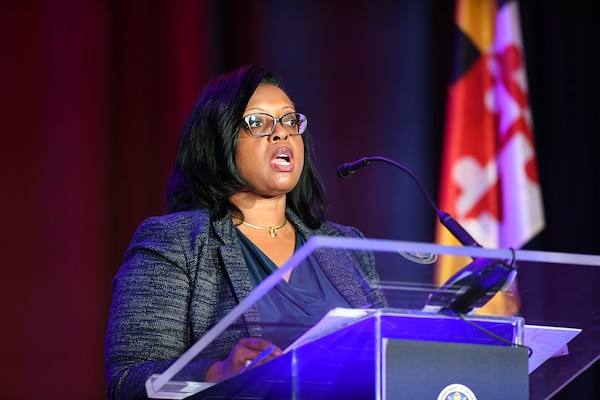 A woman wearing business attire speaks into a microphone while standing behind a lectern.