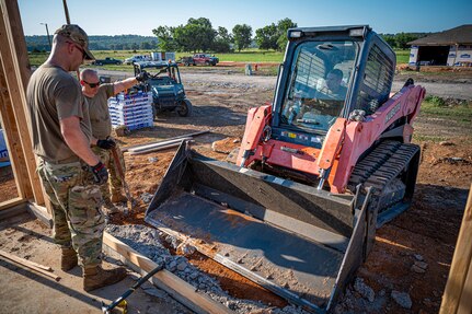 Airmen from the 103rd Airlift Wing use a skid steer to remove excess cement around the slab of a future single-family house meant for disabled veterans of Cherokee Nation in Tahlequah, Oklahoma, June 29, 2023.