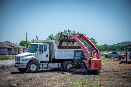 An Airmen from the 103rd Airlift Wing uses a skid steer to transfer dirt to a dump truck at the Cherokee Veterans Housing Initiative construction site in Tahlequah, Oklahoma, June 29, 2023.