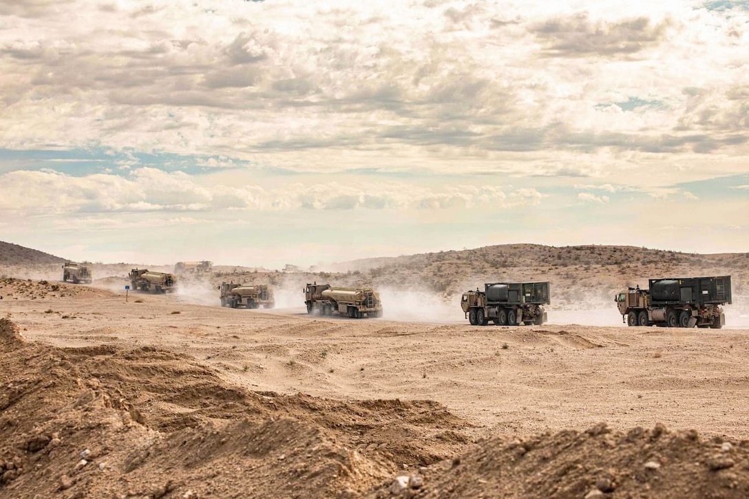 Dust fills the air as large trucks drive in formation in a desert-like area.