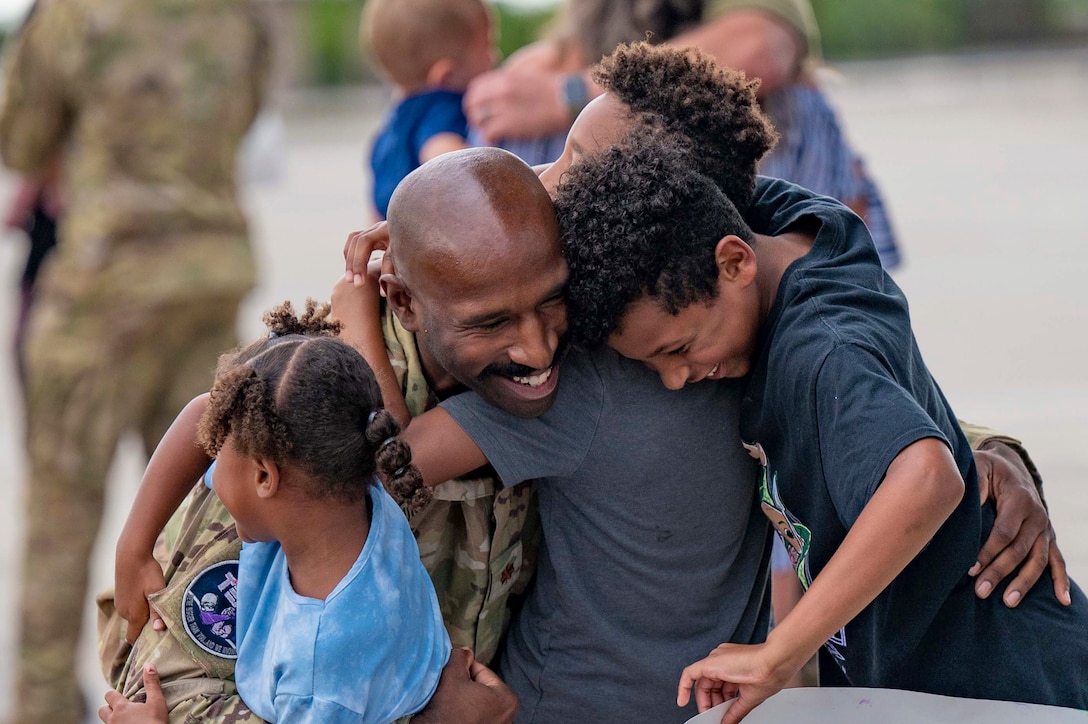 A smiling airman kneels while hugging three children.