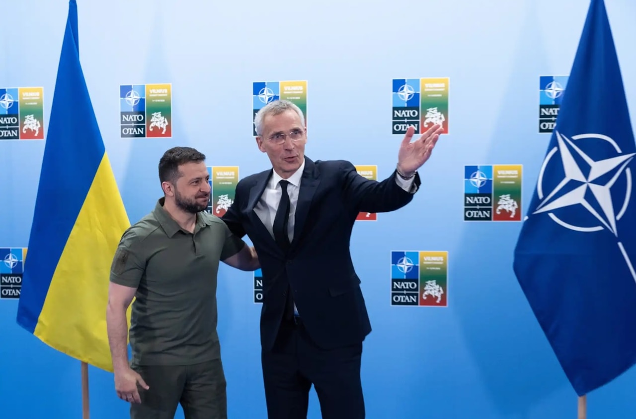 Two men stand arm-in-arm in front of flags.