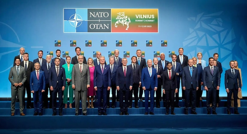 A group of people pose for a photo in front of NATO signage.