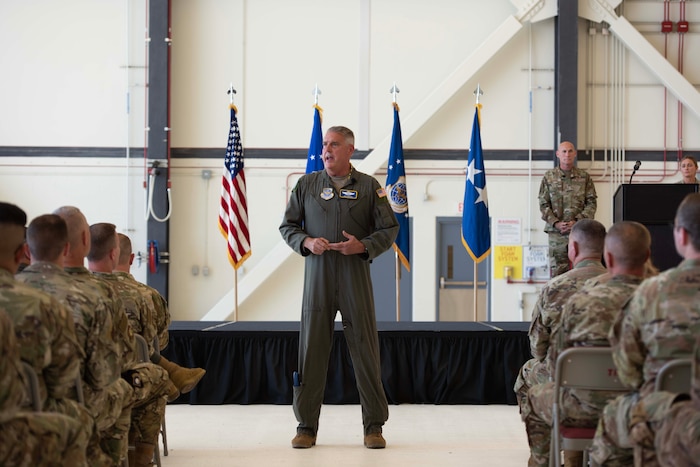 An Airman stands and speaks in front of other Airmen with a stage and flags in the background.