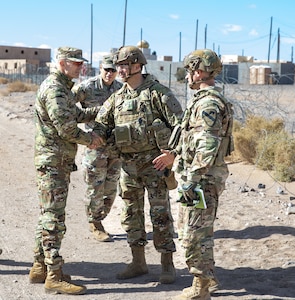 Two men in military uniforms shake hands. Other men in uniform stand nearby.