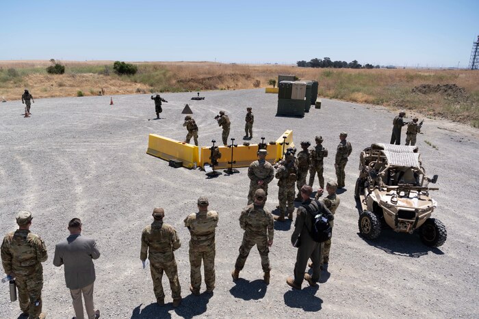 Airmen gather around an obstacle course where other Airmen demonstrate a training exercise.