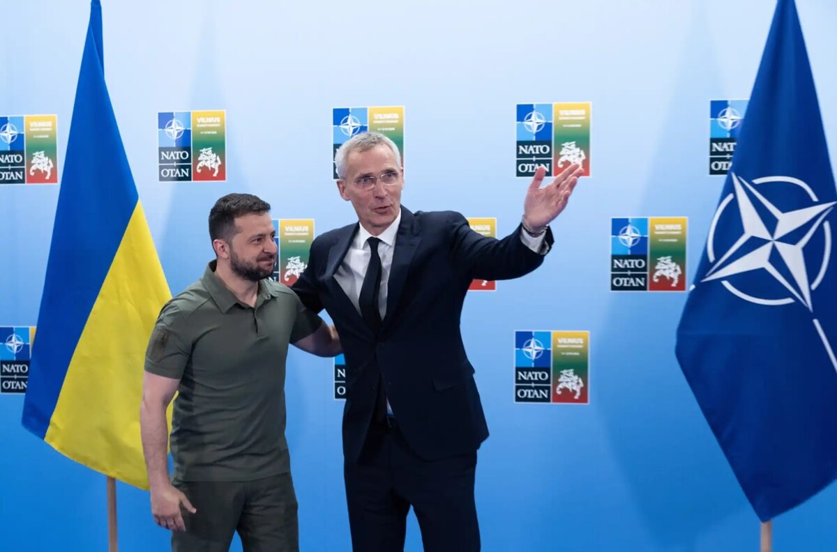 Two men stand arm-in-arm in front of flags.
