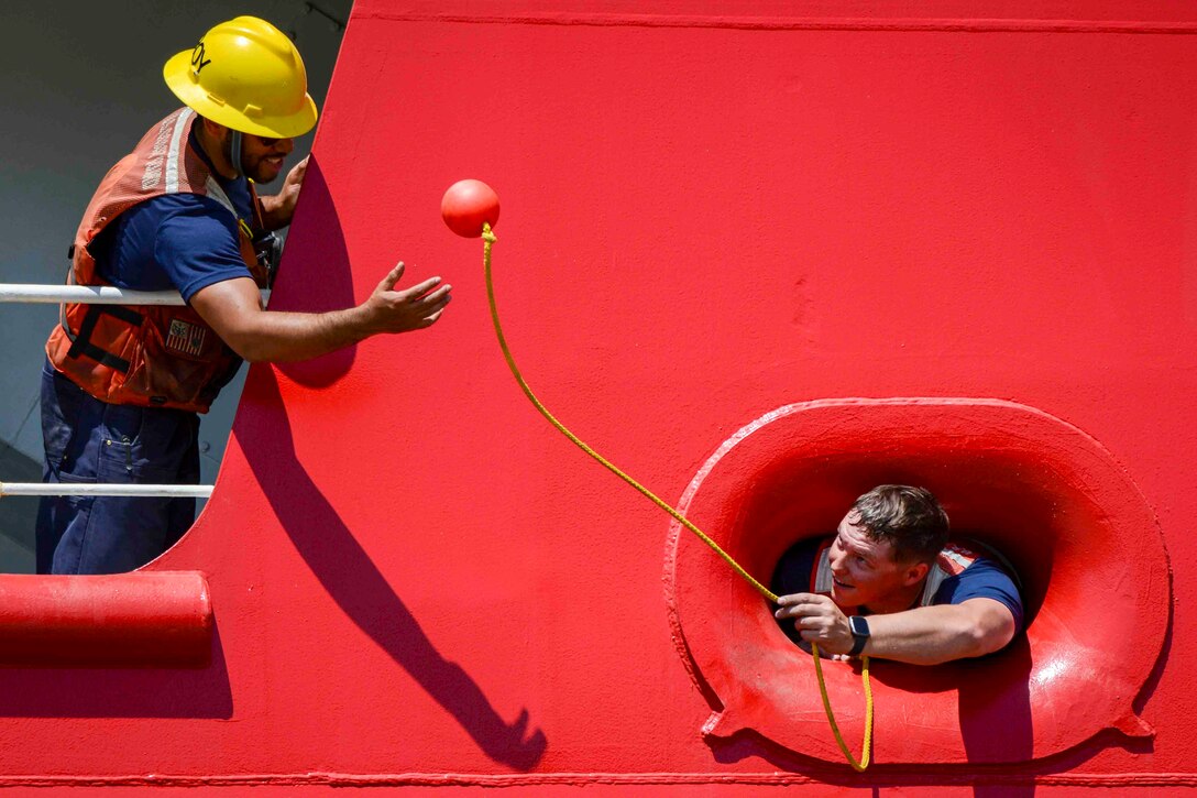 A Coast Guardsman tosses a heaving line to another Coast Guardsman from the porthole of a ship.