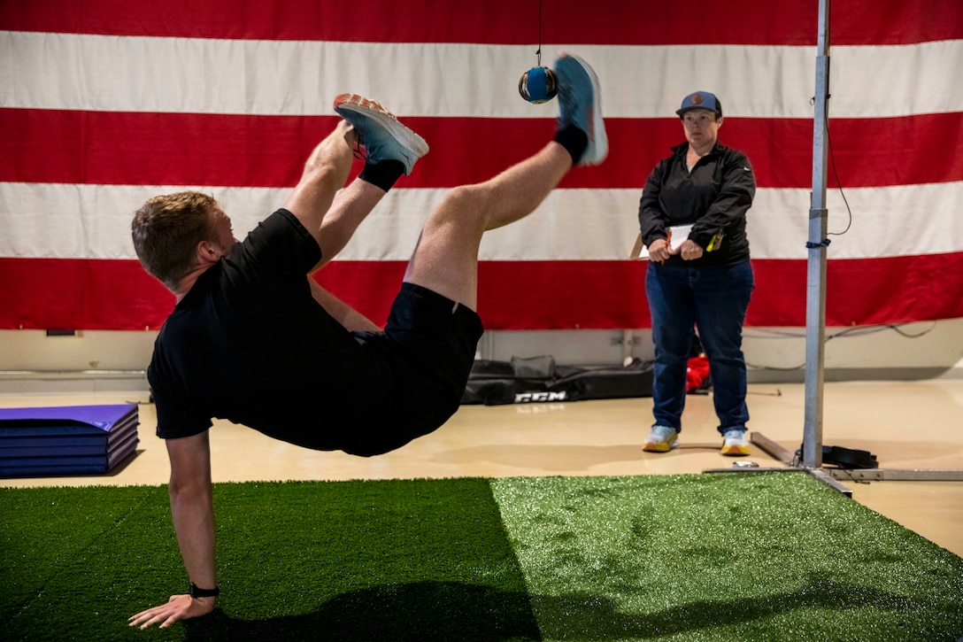 A guardsman performs high kicks on a turf as a judge evaluates while holding a clipboard.