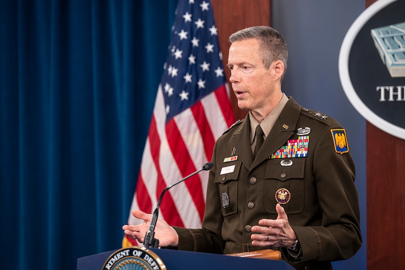 A uniformed service member stands at a podium with flags in the background.