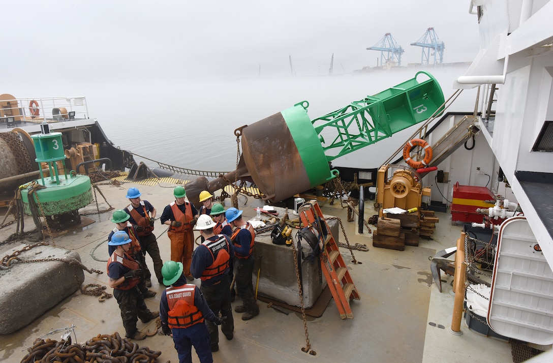 A group of people with blue, green and whit hard and orange safety vest stand next to a large green and grey concrete and steel bouy.