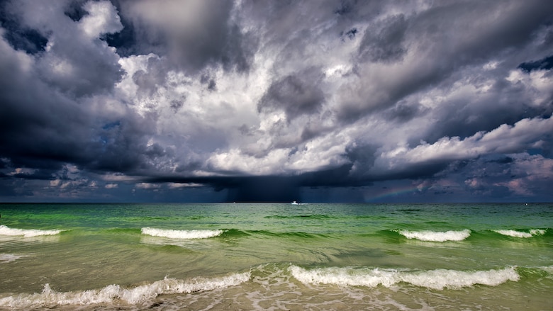Large grey storm clouds are show over a large body of water, the Gulf of Mexico, the water is reflecting the colors red, green and blue making it look like the beginnings of a rainbow.