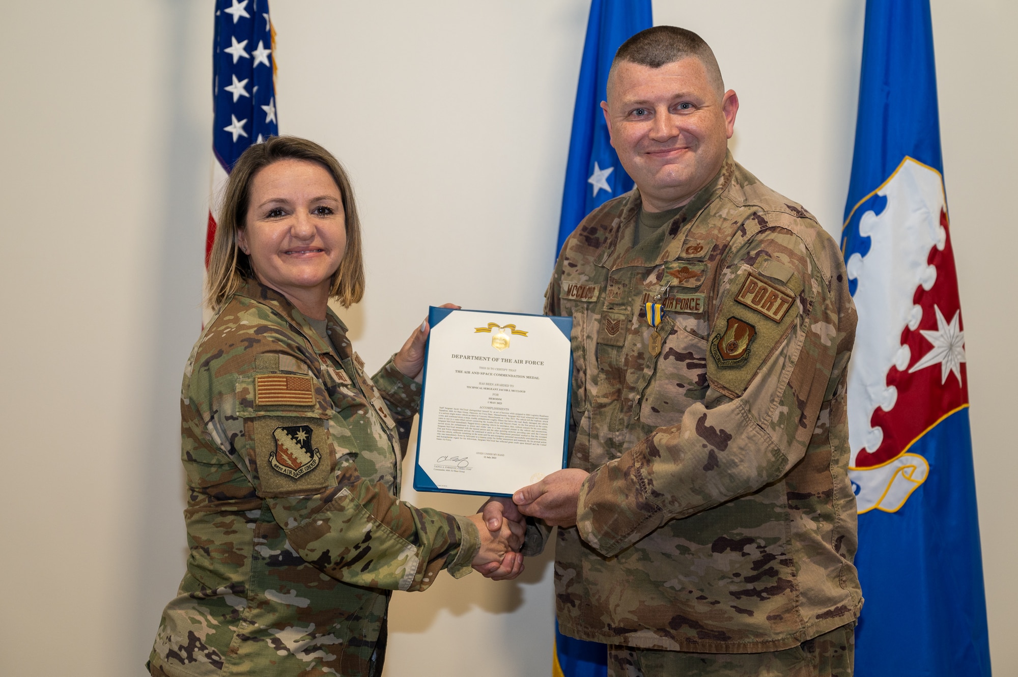 two uniformed armed pose in front of the American flag; the colonel presents the tech sergeant with a medal and printed orders.