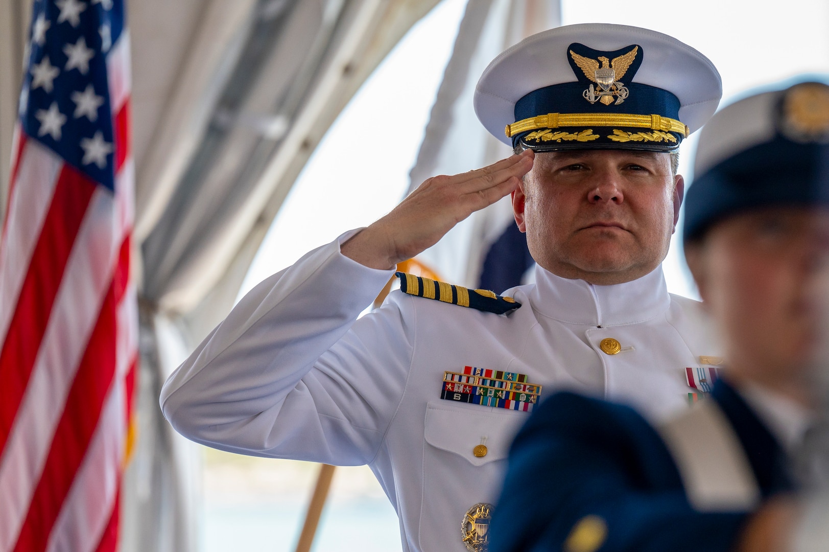 officer in dress whites salutes, flag in background