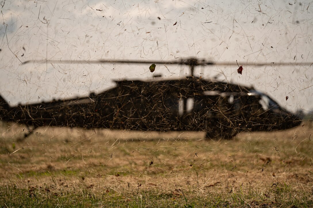 Debris is blown into the air as a helicopter lands on a grassy area.