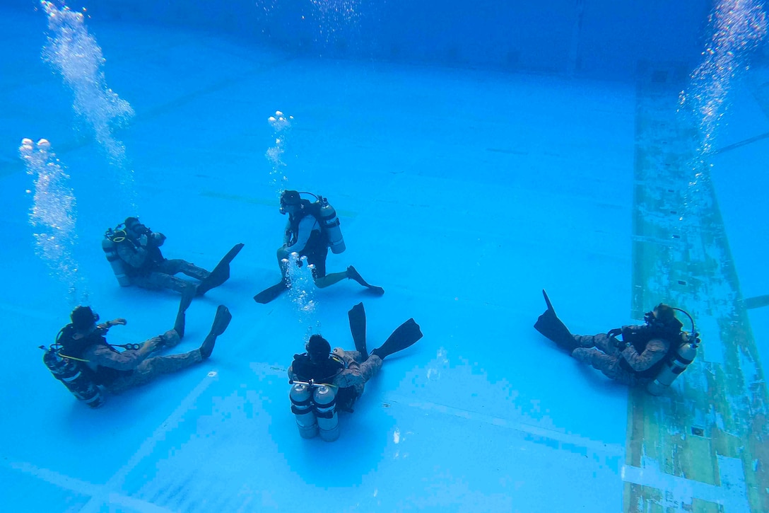 Five Marines wearing dive gear sit underwater at the bottom of a pool.