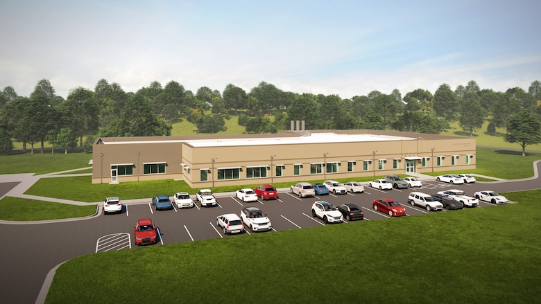 A tan building with cars parked in front, grass on all sides, and trees in the background.