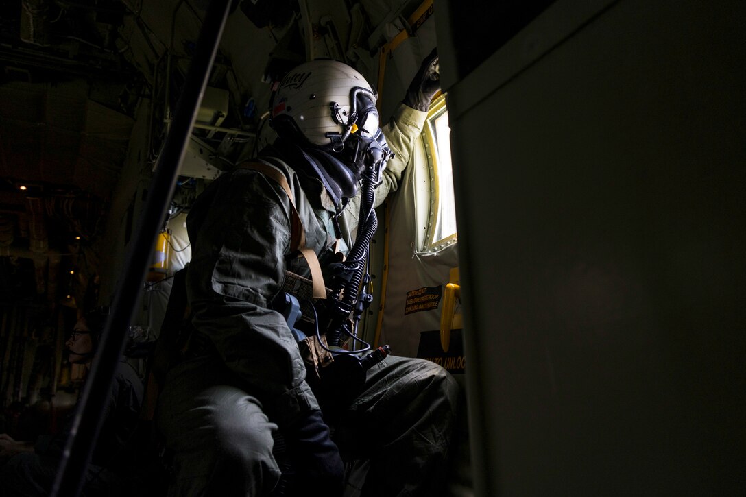 A Marine wearing protective gear looks out of the window of an aircraft.