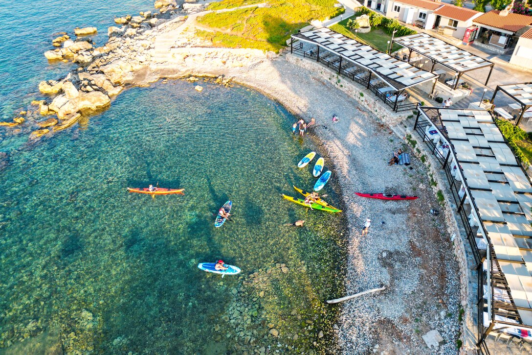 People ride in colorful kayaks at a rocky beach as a shelter covers table and chairs to the right.