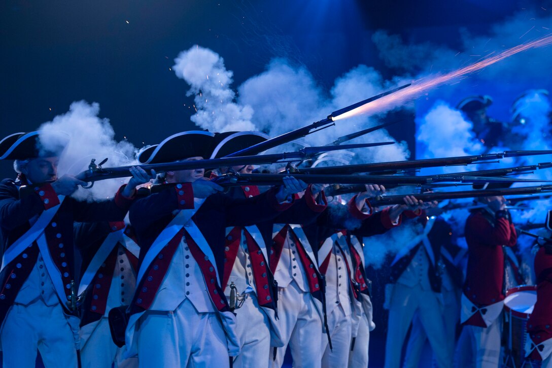 Smoke surrounds soldiers dressed in ceremonial uniforms as they fire weapons covered in a blue haze.