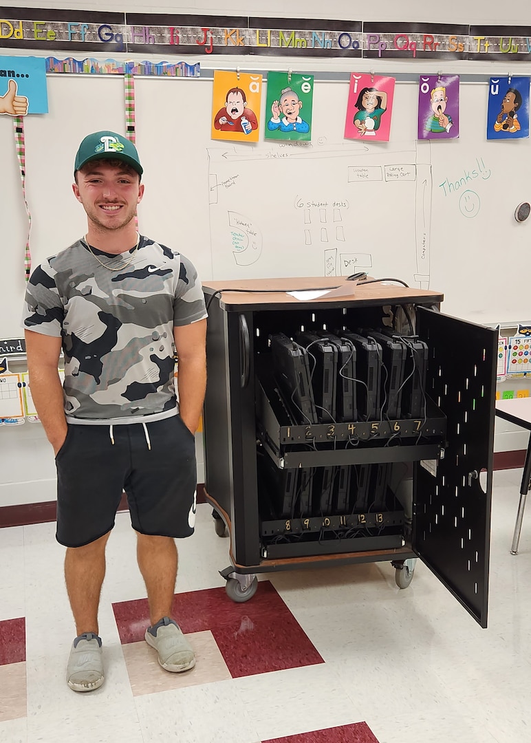 A young man stands next to a cart on wheels that is almost as tall as his shoulders. He has his hands in his shorts pockets, a gray camo shirt on and a green ball cap.
