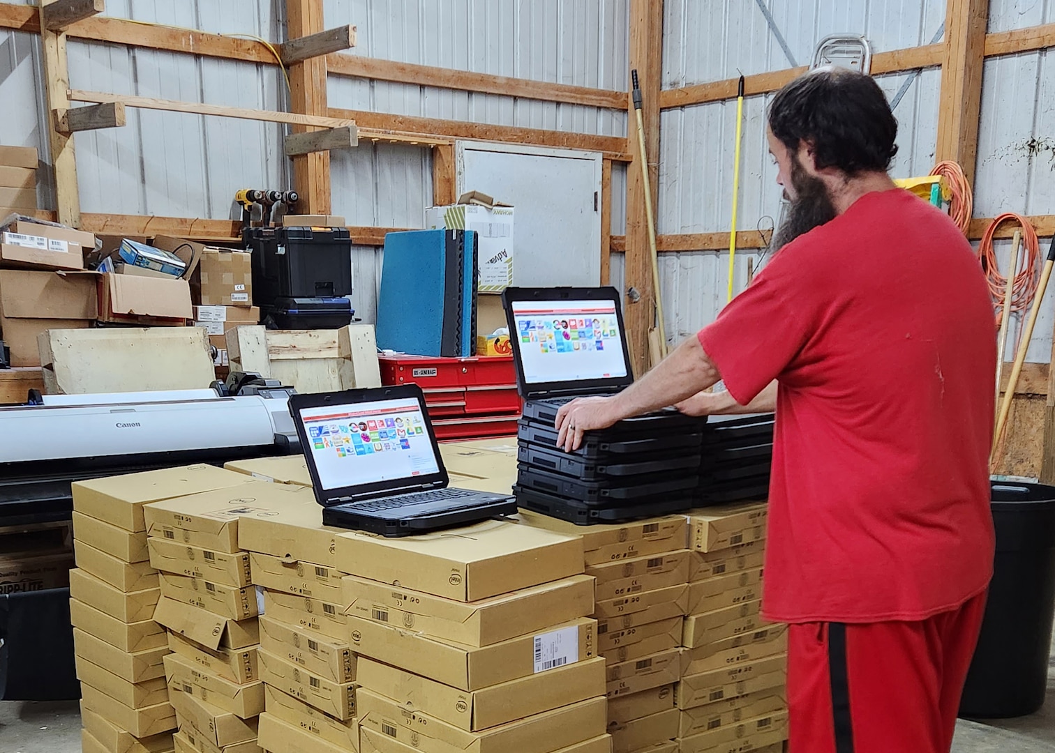A man in a red shirt with his back to the camera stands looking at two operating and open laptop computers. They are setting on a large stack of brown boxes that look like they may contain laptops as well.