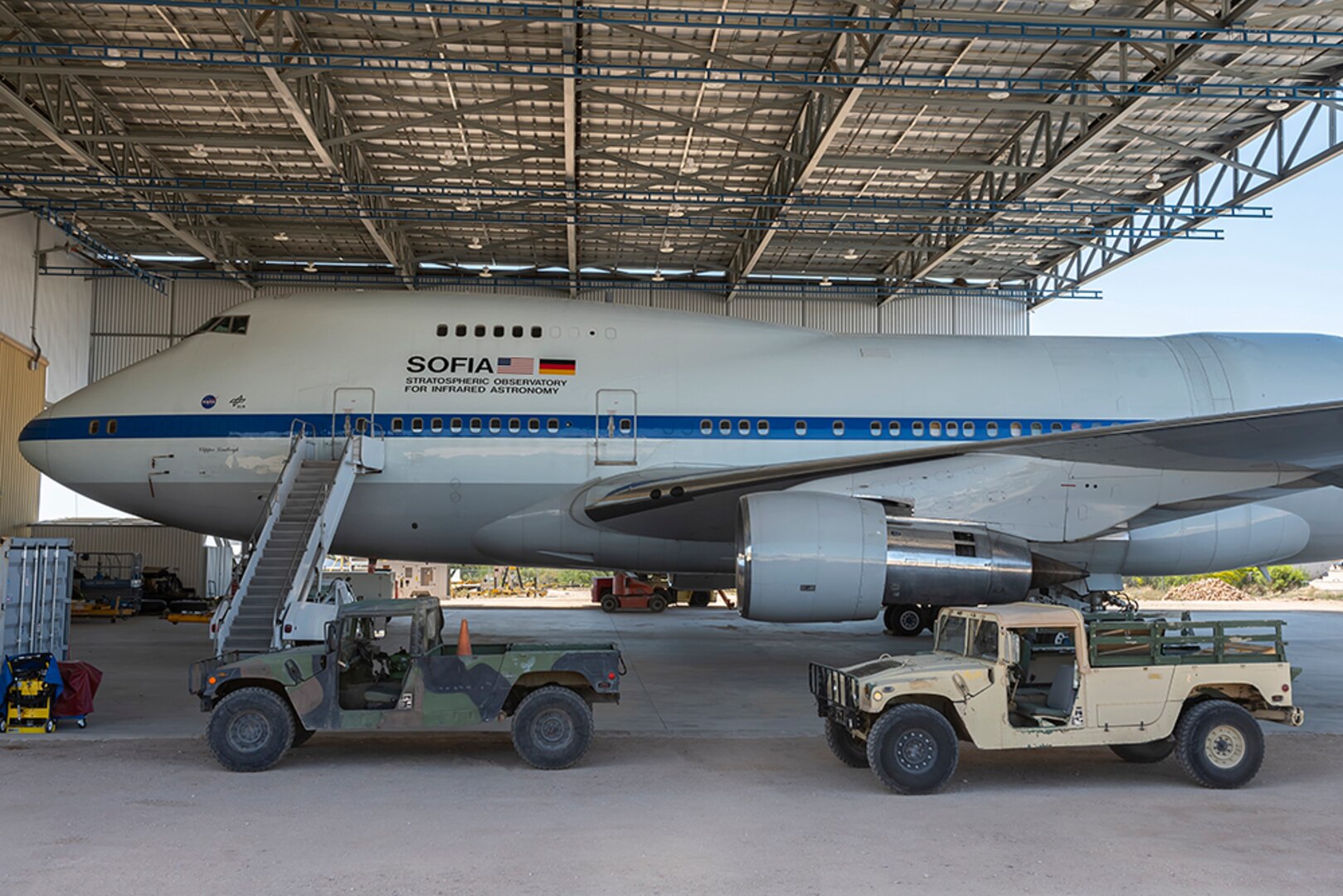 A Boeing 747 and a Humvee are parked in a hangar in the desert.