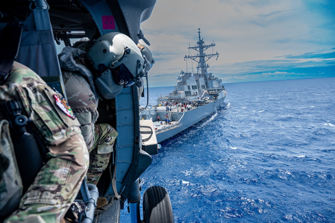 Service members in a helicopter fly over water as a Navy ship sails behind them.