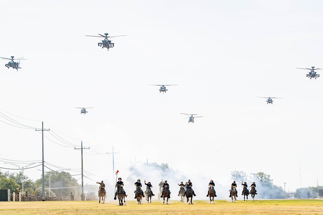 Soldiers ride horses across a field as military aircraft hover above them.