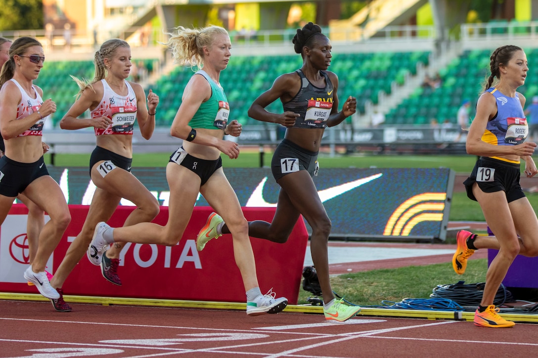 A soldier runs with other competitors in a stadium with bleachers in the background.