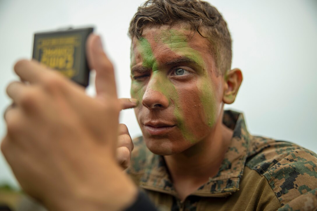 A close-up of a Marine shutting one eye while holding a compact as he applies camouflage to his face.