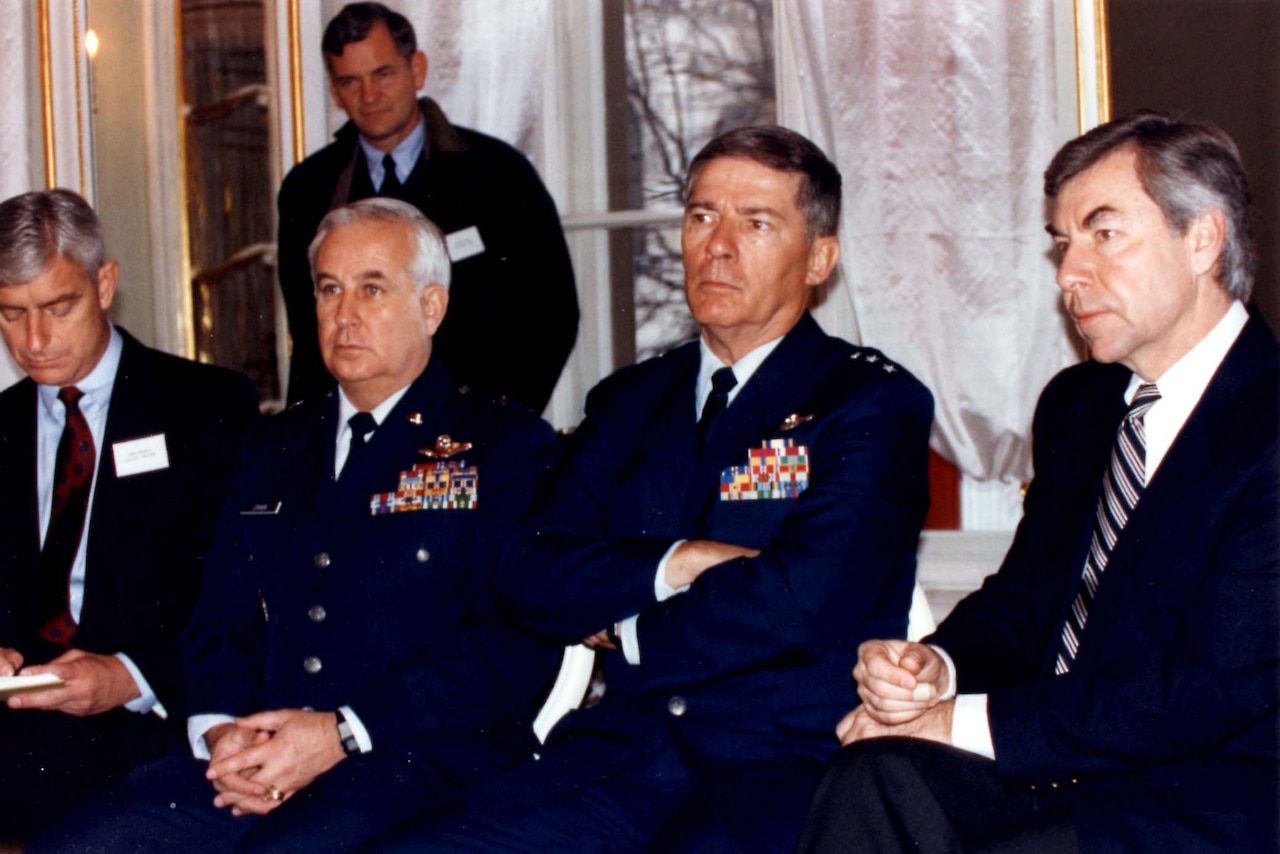 Four men in military dress uniform are seated. Another man stands in the background.
