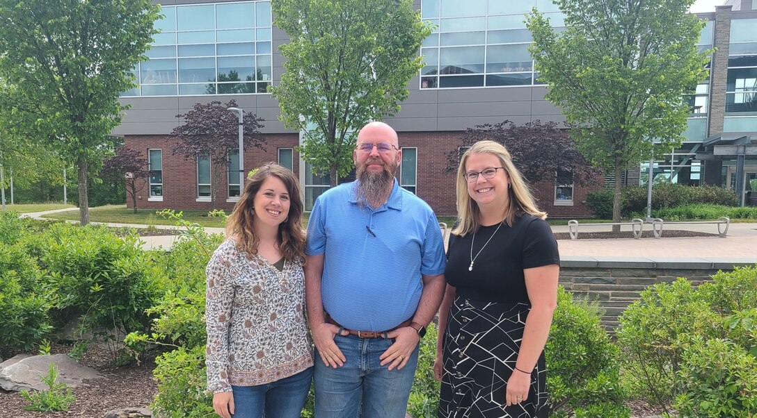 The image depicts three people standing outdoors posing for a photo.  They are smiling.  Behind them, a building and trees can be seen.