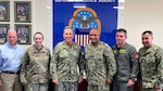 Five men and one woman stand in front of a picture of the DLA logo