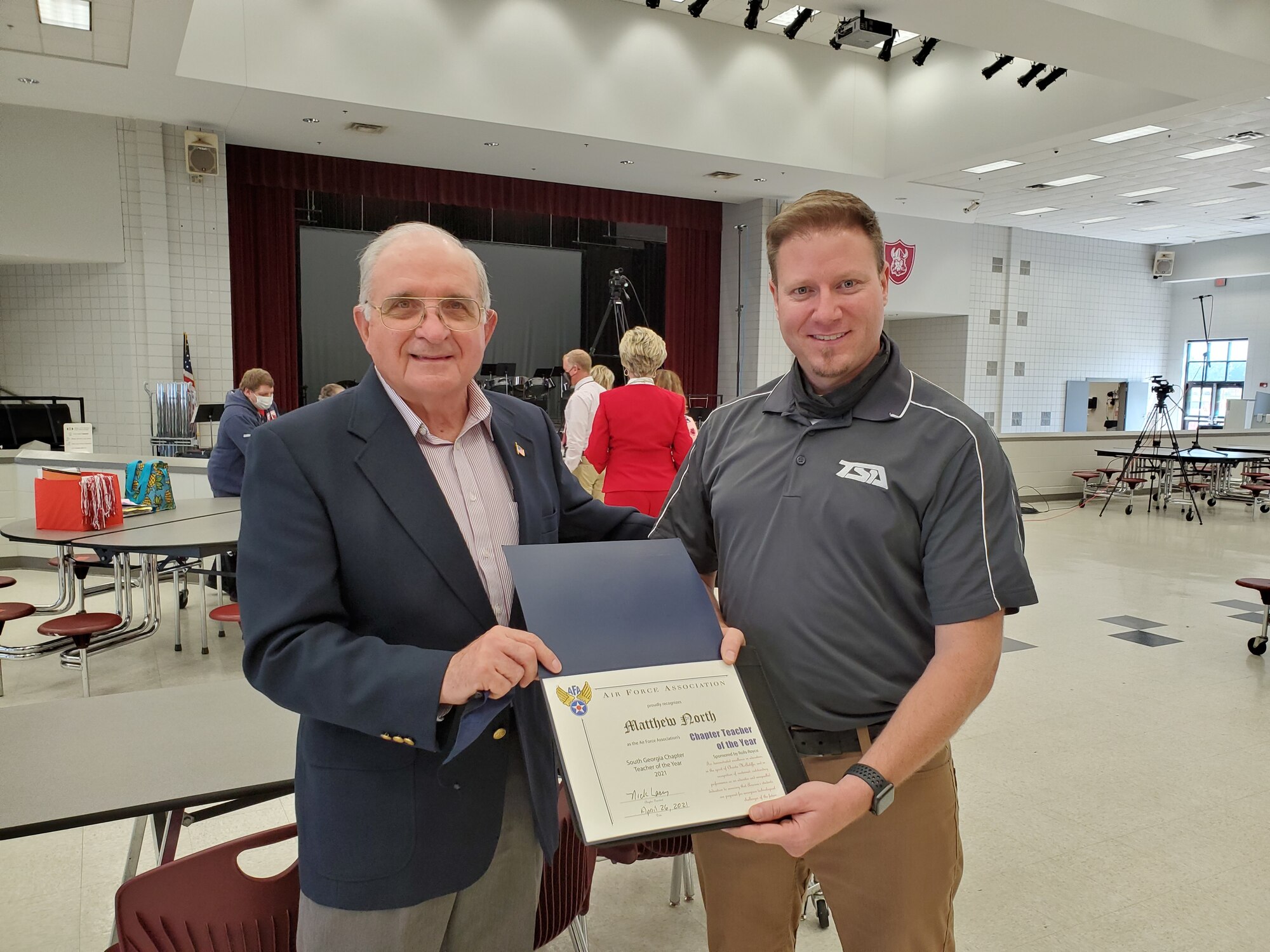 A photo of two people holding a certificate between them in a school cafeteria.