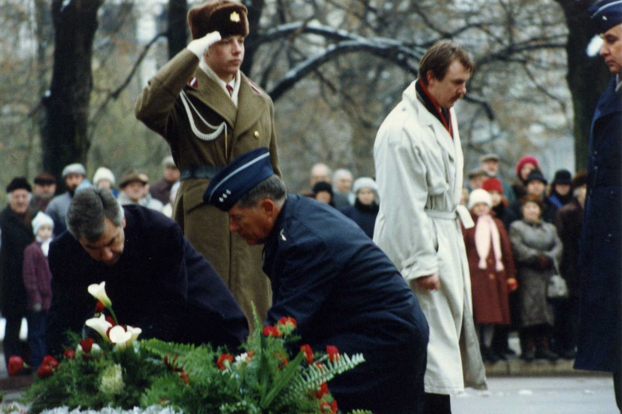 A foreign troop renders a salute while two men in military uniform lay a wreath.