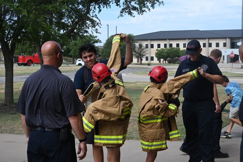 Firefighters assigned to the 17th Civil Engineer Squadron assist children with putting on gear for a simulated search and rescue exercise during Junior Firefighter Camp at the fire station, Goodfellow Air Force Base, Texas, July 12, 2023. Junior Firefighter Camp puts the children in simulated training environments to teach basic firefighting skills in an engaging way. (U.S. Air Force photo by Airman 1st Class Madison Collier)