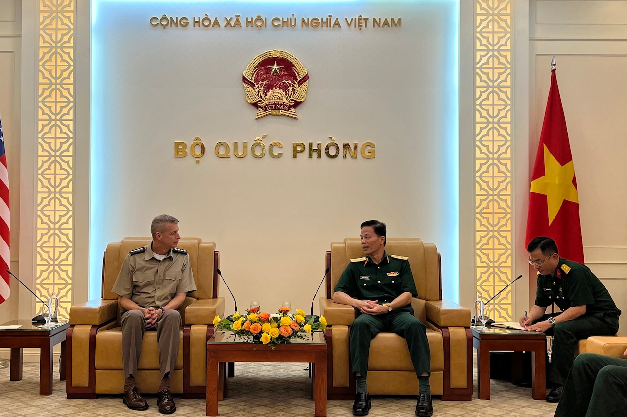 Two men in uniform sitting down in chairs talk with each other next to the United States and Vietnam flags. One man in uniform takes notes.