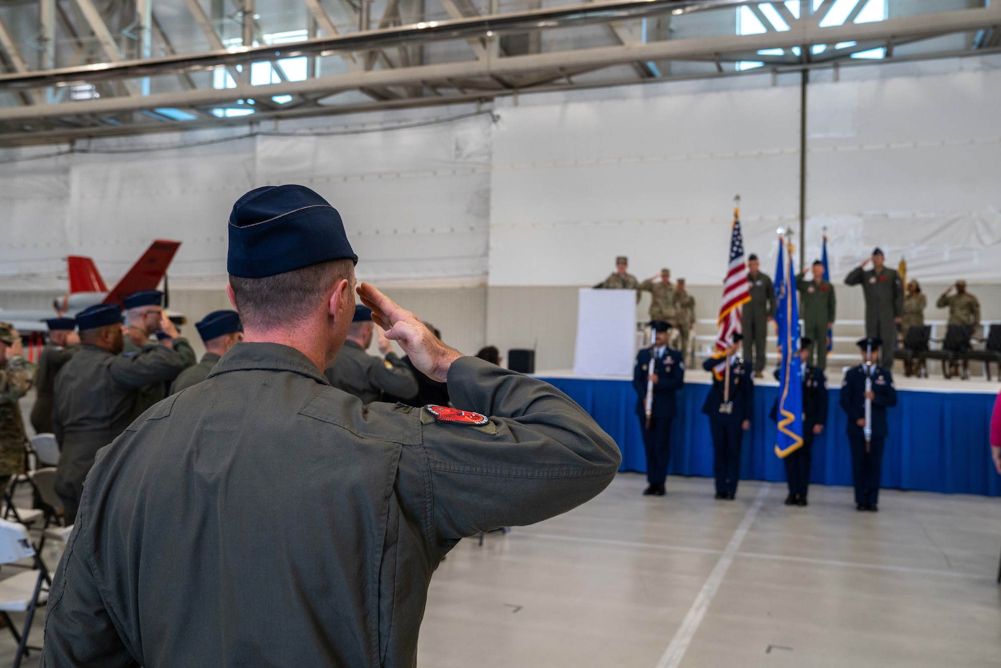 A man in a military flight suit salutes before an American flag.
