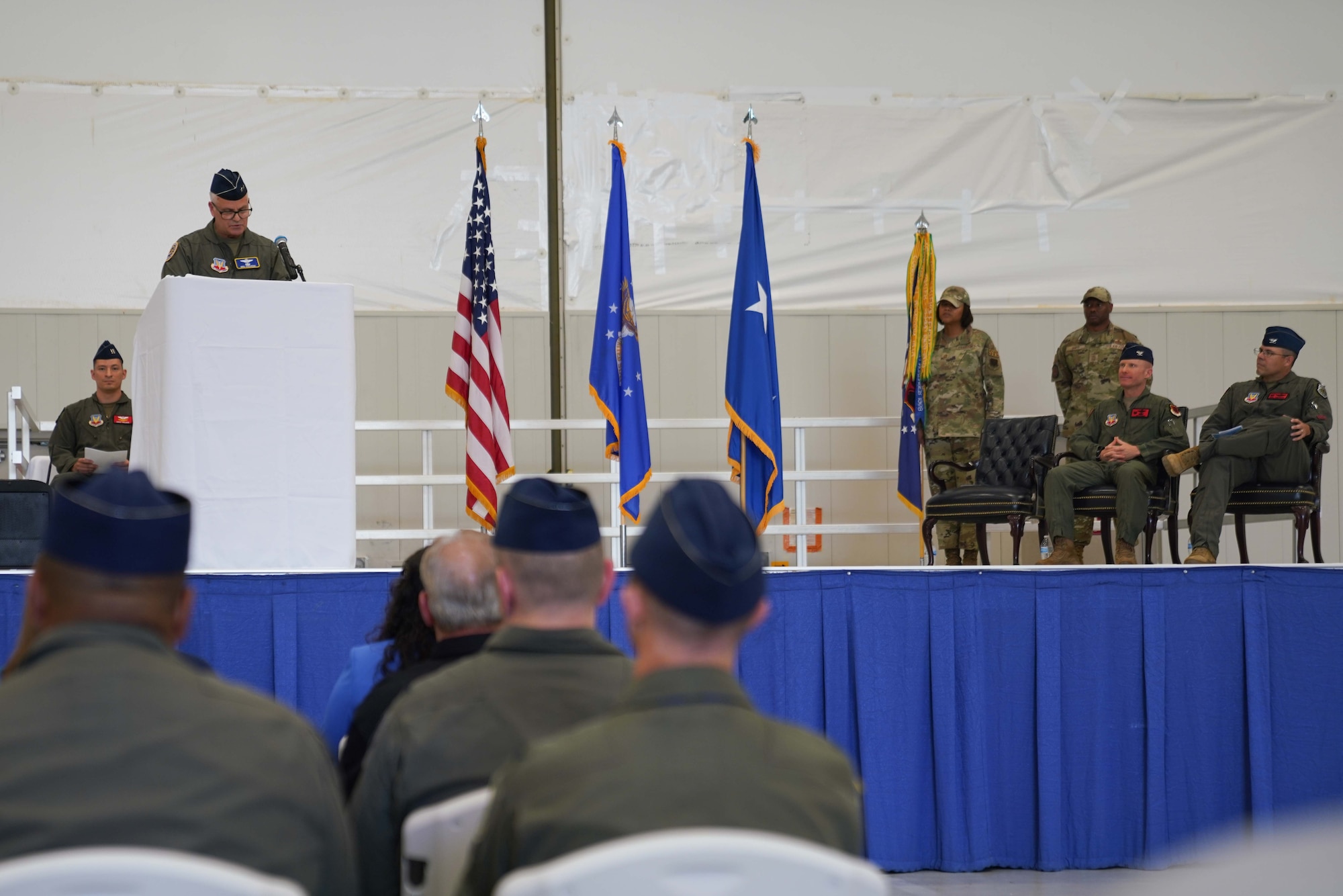 Military men in flight suits sit on a stage while another man in a flight suit speaks at a podium.
