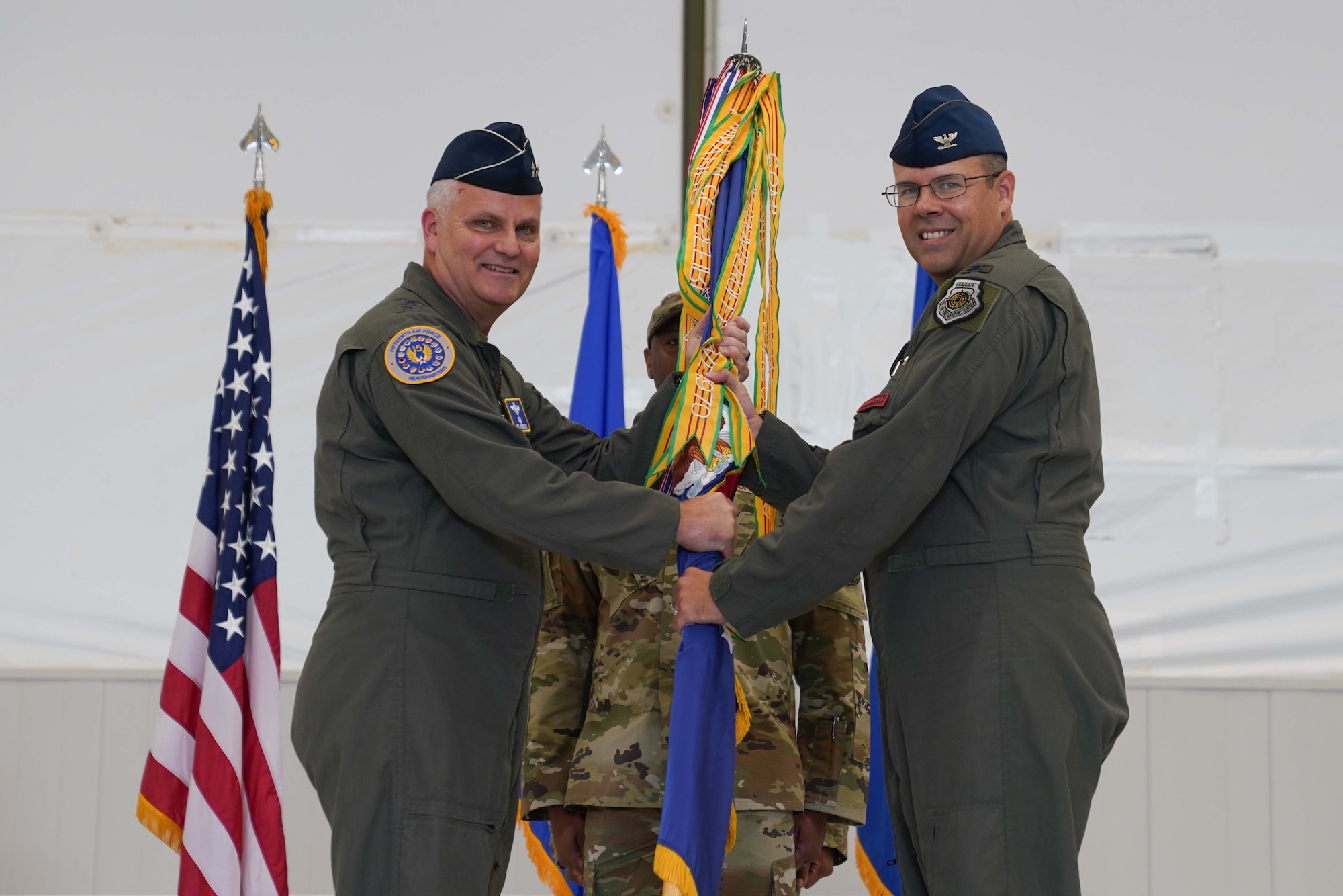 Two men in military flight suits hold a pole with a pennant on it and smile.