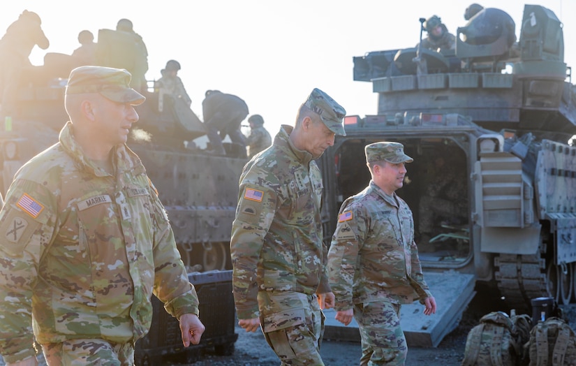 Three men walk past military equipment.