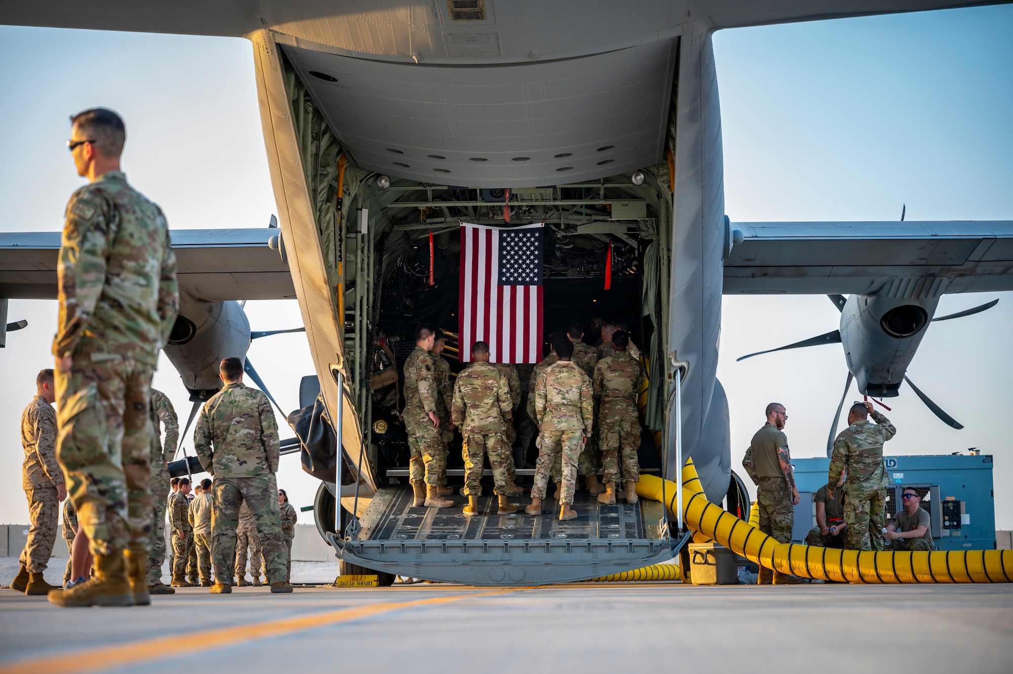 An American Flag hangs over military members in uniform, as they stand on the back ramp of an aircraft waiting to tour the inside.
