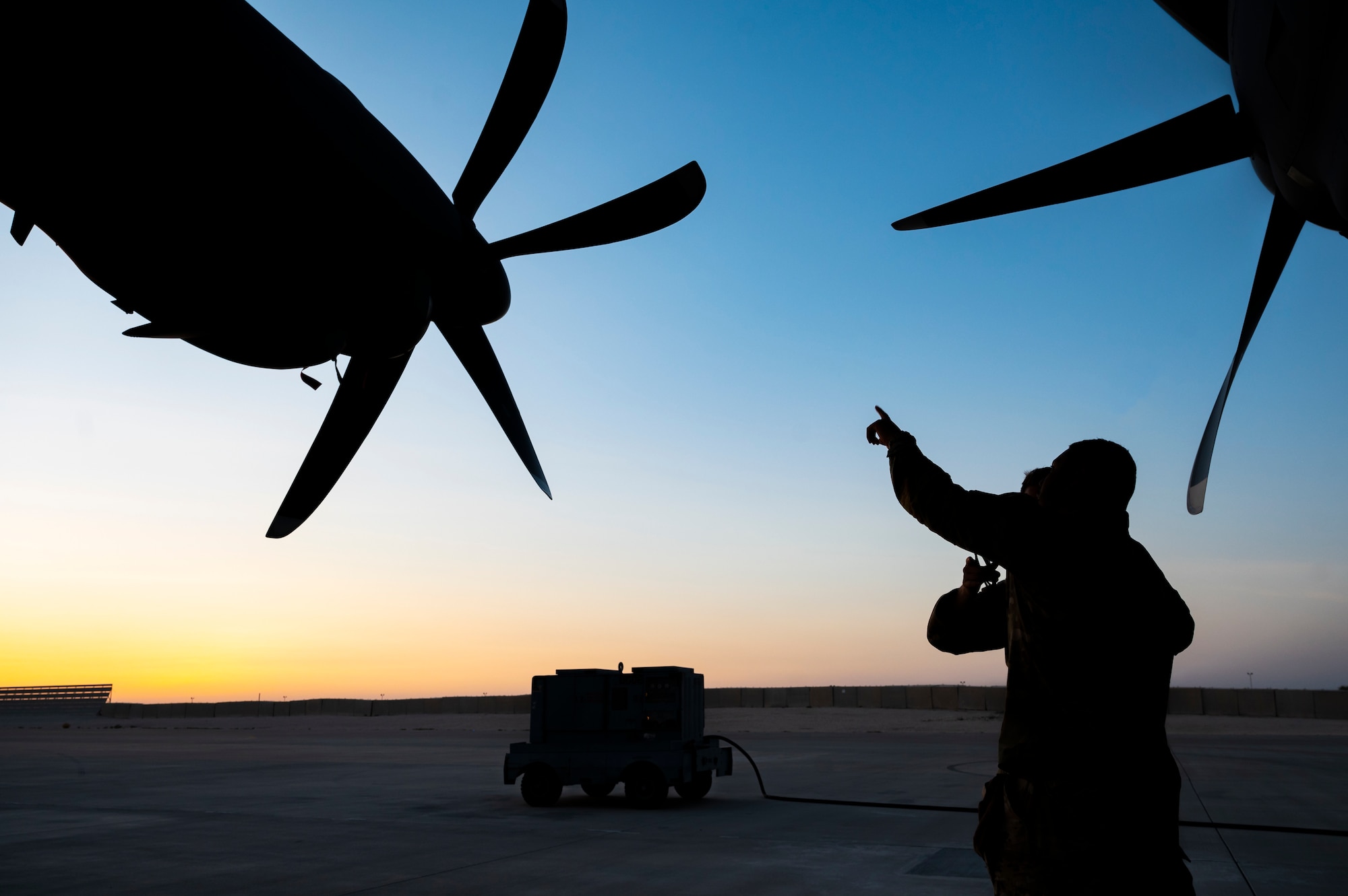 Military members in uniform point at the engine of an aircraft, while standing underneath the wing.