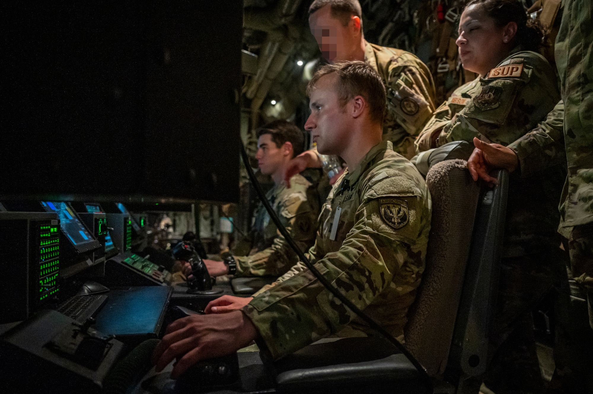 Military members in uniform sit and use a joystick to control cameras attached to an aircraft.