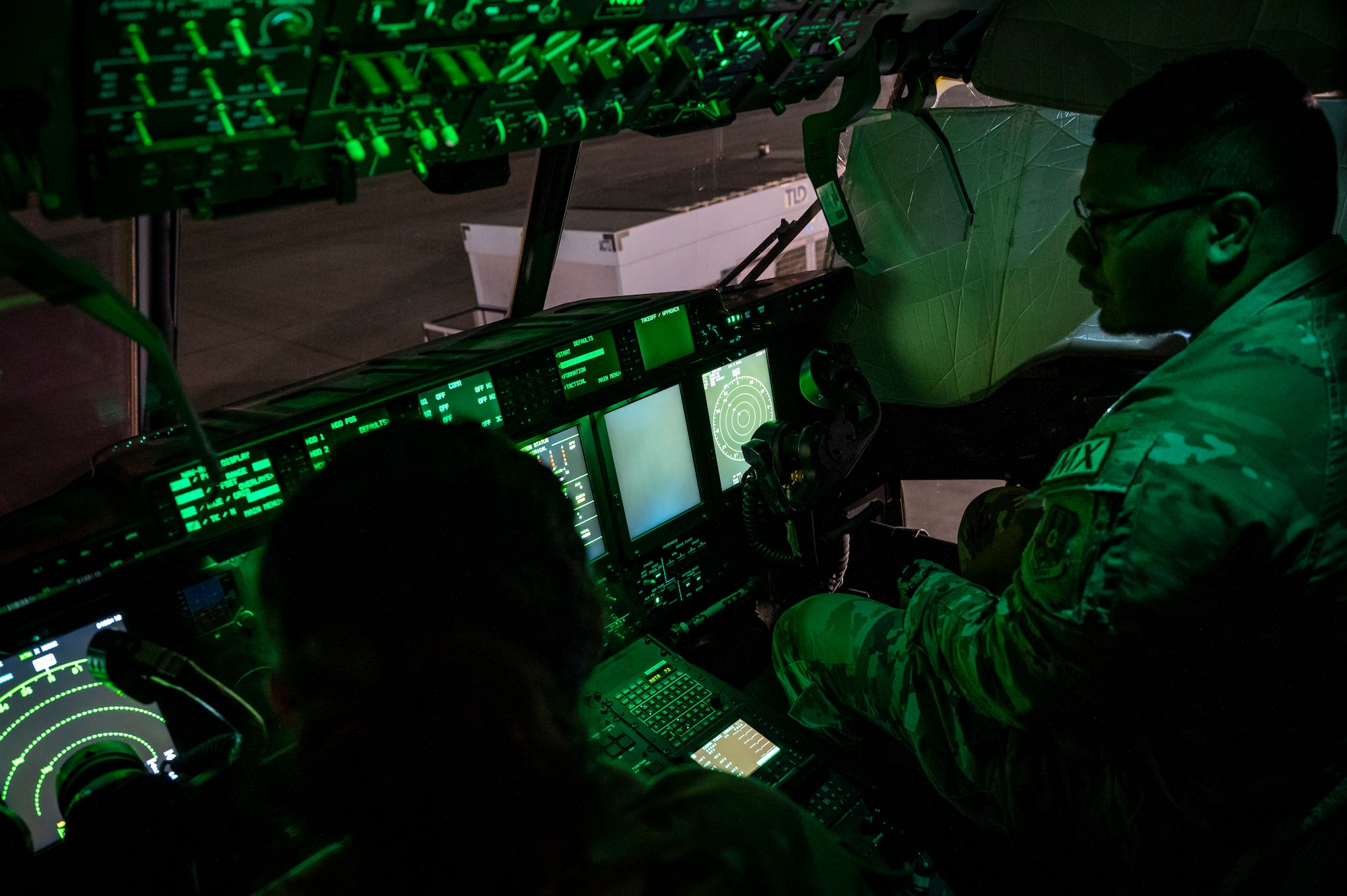Two military members in uniform sit in the pilot and co-pilot seat of an aircraft.